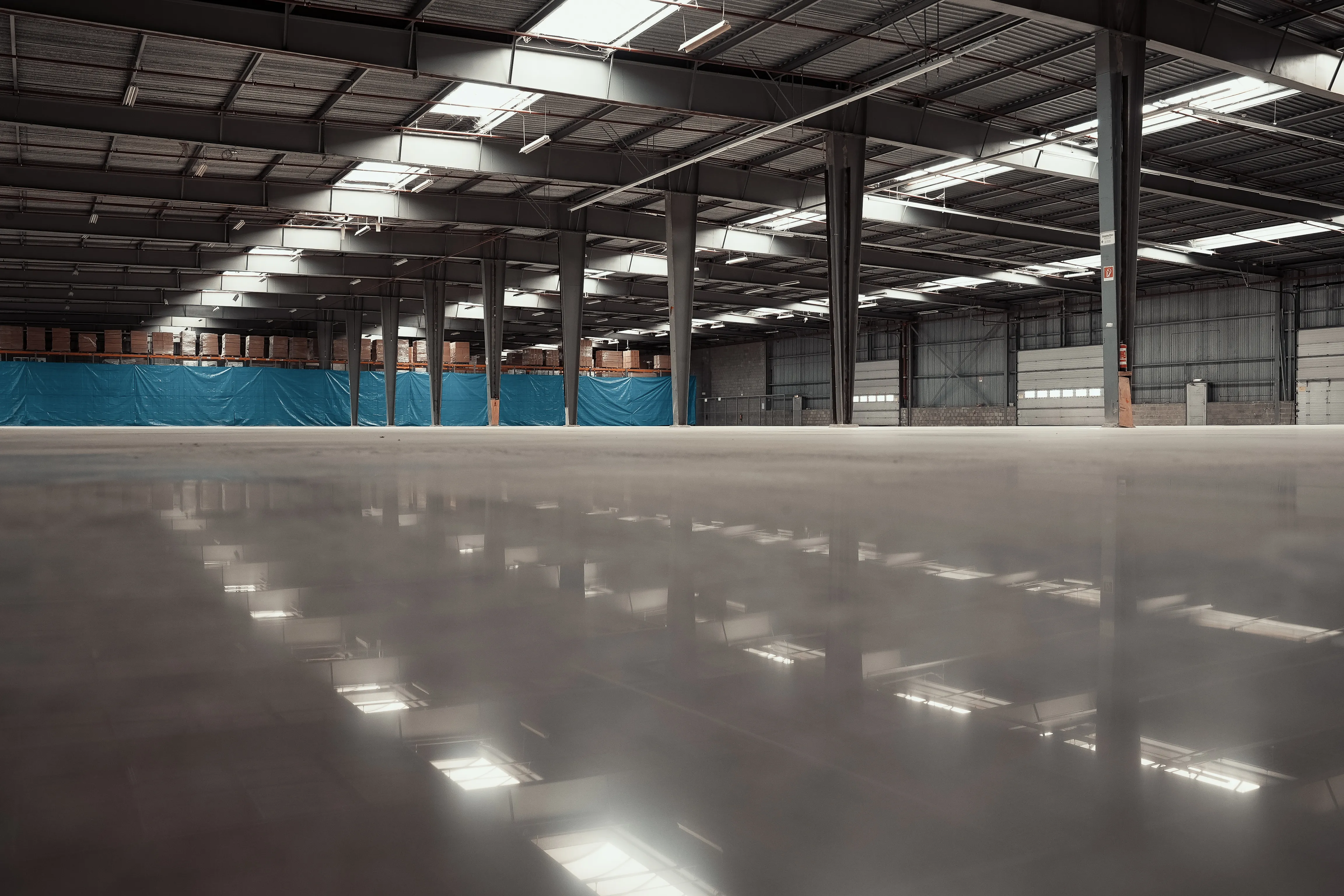 Groot magazijn met een gepolijste betonvloer die zo glanst dat de lichten en de dakconstructie erin weerspiegelen. Op de achtergrond stalen kolommen, rekken met pallets en een blauw afdekzeil.