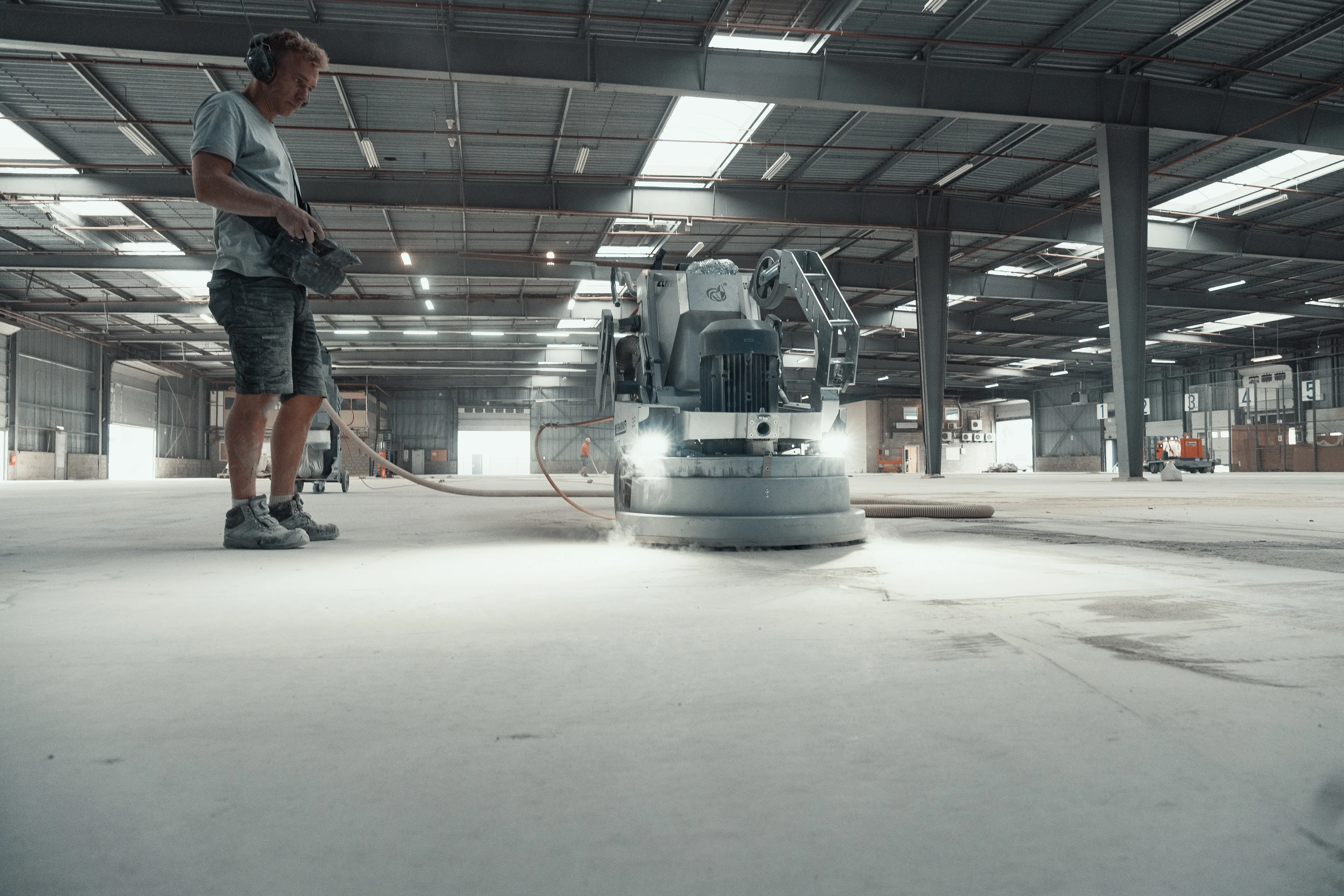Medewerker met gehoorbescherming bedient een grote vloerschuurmachine in een lege industriële hal. Onder de schijf stuift betonstof op, met slangen naar een stofafzuiging op de achtergrond.